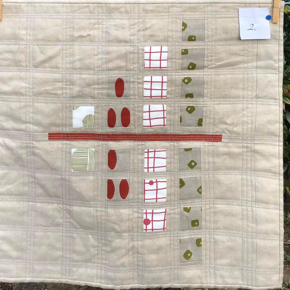 A square quilt with a beige background. The quilt features a symmetrical pattern of red circles, green squares, and beige shapes, arranged in rows. A red line divides the quilt horizontally.