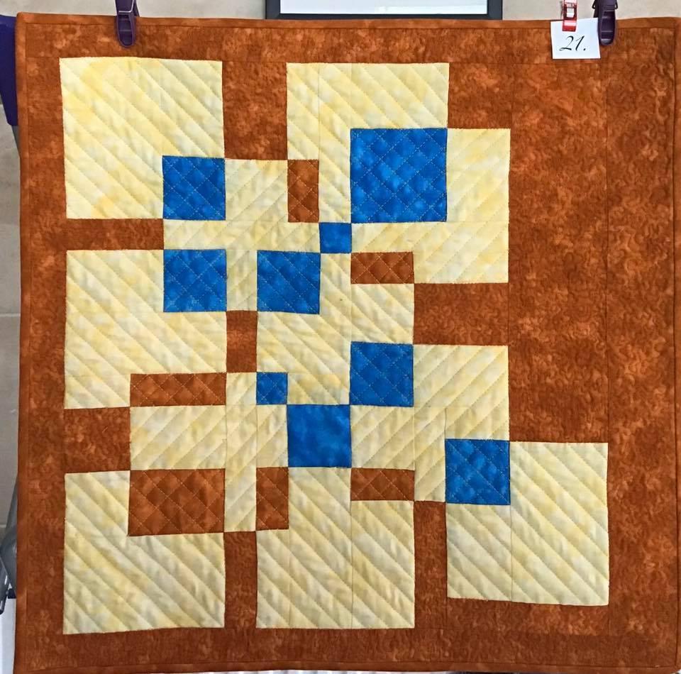 A small, square quilt with a brown background. The quilt features a pattern of orange and blue squares of varying sizes, arranged in a seemingly random pattern. The background fabric has a subtle, textured pattern.
