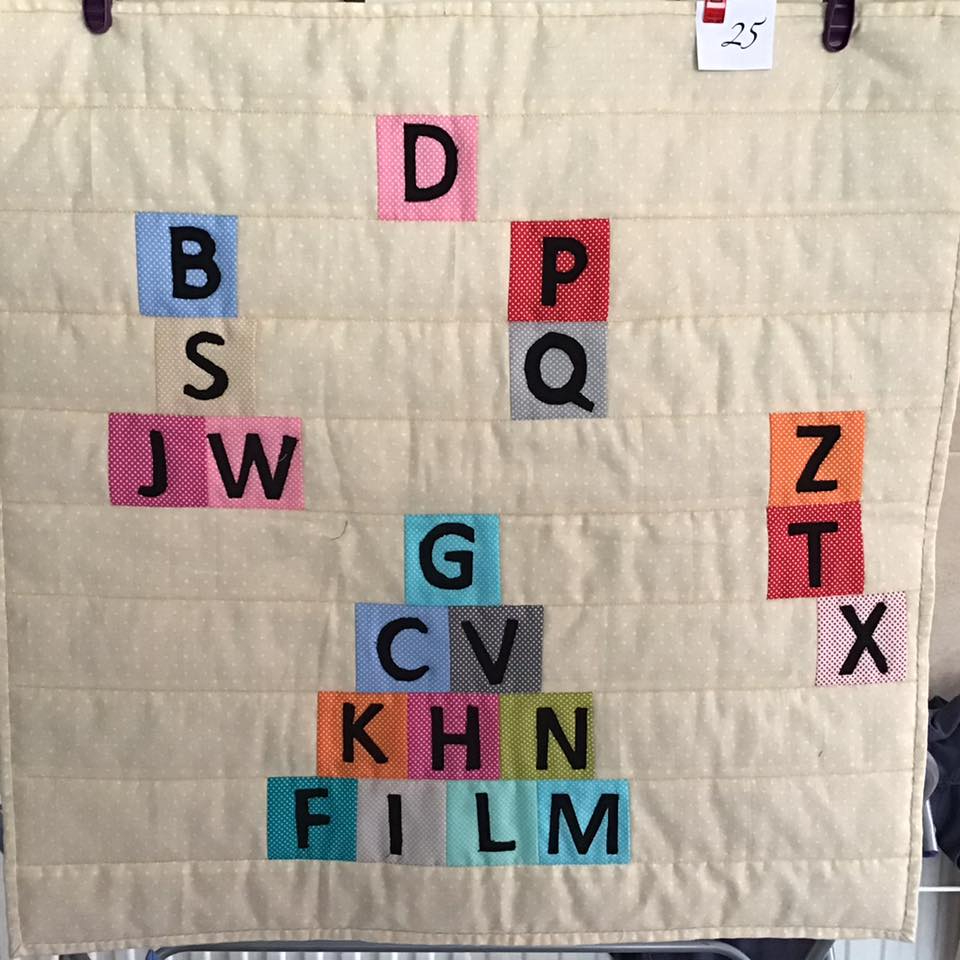 A small, square quilt with a beige background. The quilt features letters of the alphabet arranged in various patterns, including a pyramid shape and a vertical line. The letters are colourful and sewn onto the fabric.