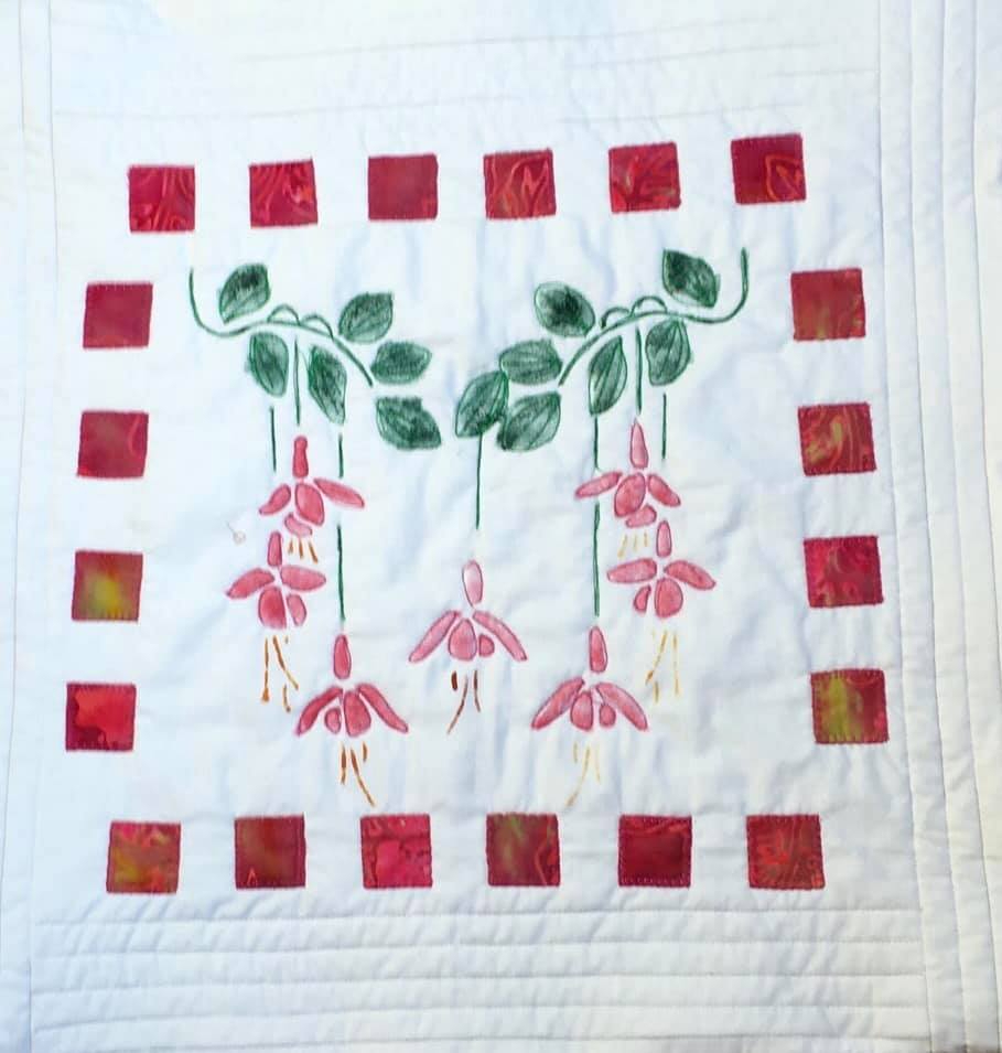 A small, rectangular quilt with a white background. The quilt features a row of pink fuchsia flowers hanging from green vines. The flowers are surrounded by a border of red squares.