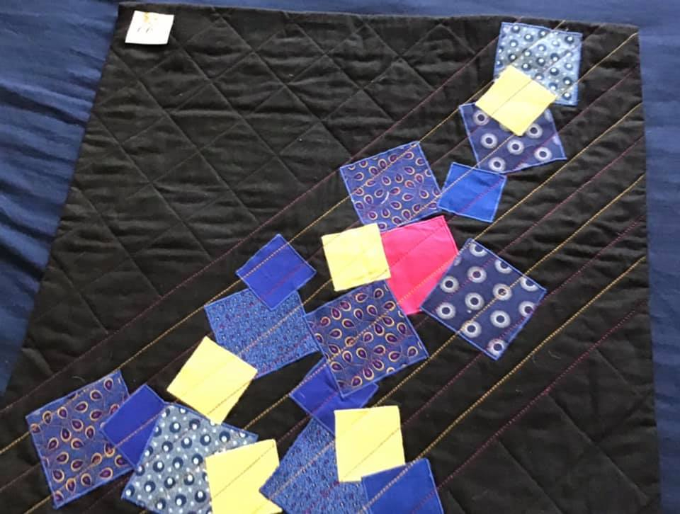 A small, square quilt with a black background. The quilt features a diagonal line of colourful squares in blue, yellow, and pink. The squares are made of fabric with different patterns, including polka dots and paisley. The background is quilted with a diamond pattern.