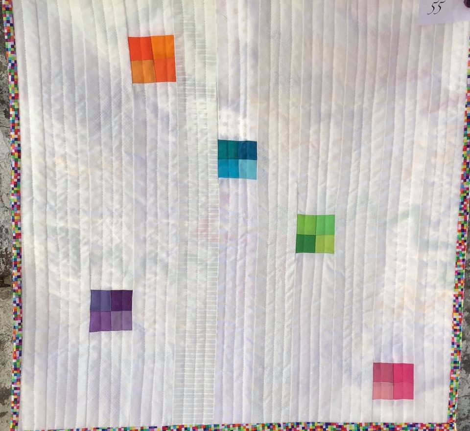 A small, square quilt with a white background. The quilt features five colourful squares of different sizes, each made up of smaller squares in a gradient of colours. The background is quilted with vertical lines.