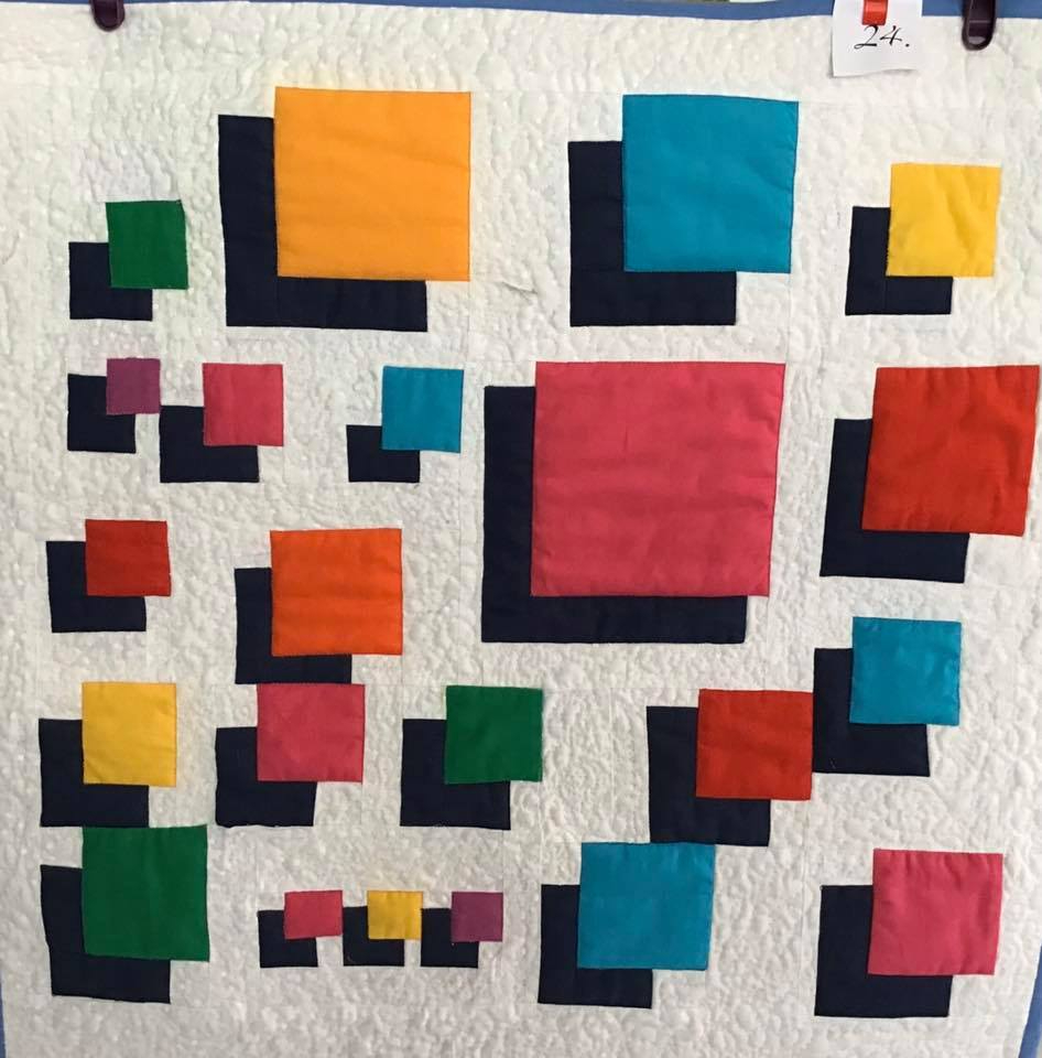 A small, square quilt with a white background. The quilt features a pattern of colourful squares in various sizes and colours, including red, pink, yellow, green, and blue. Some squares overlap each other, creating a layered effect.