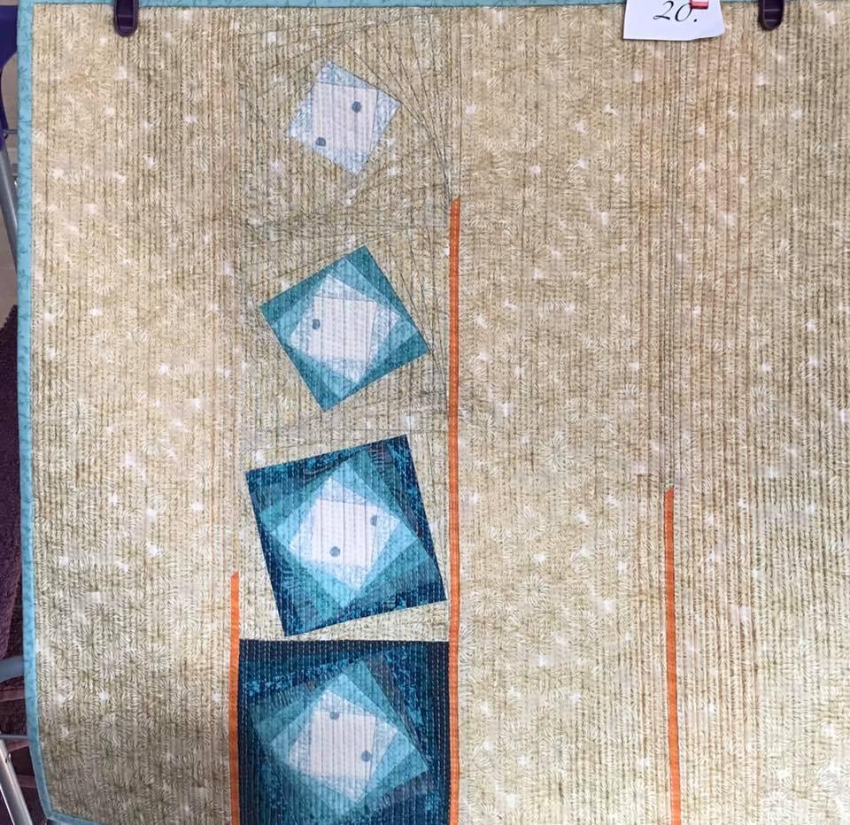 A small, square quilt with a beige background. The quilt features a pattern of blue squares and rectangles arranged in a vertical line, with orange lines separating them. The background has a subtle, textured pattern.