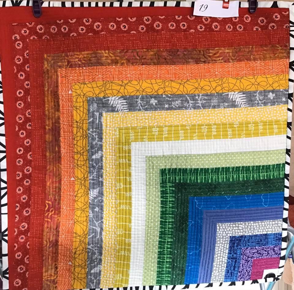 A small, square quilt with a rainbow-inspired design. It features strips of fabric in various colours, from red to purple, arranged in a diagonal pattern. Each strip has a different texture or pattern.