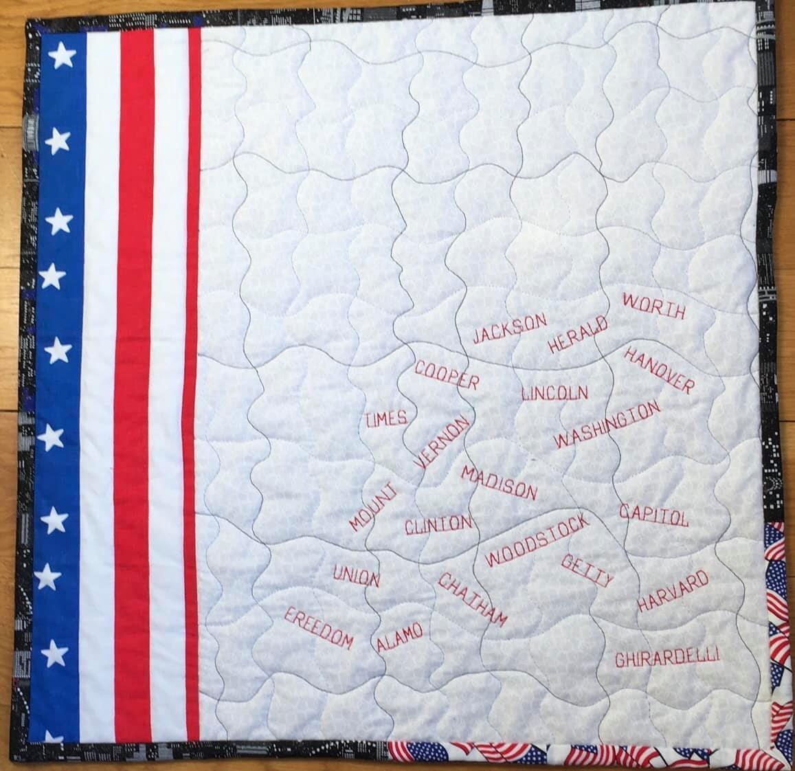A quilt in the shape of the United States, with each state outlined and labeled. The quilt has a patriotic colour scheme of red, white, and blue, with stars along the sides.
