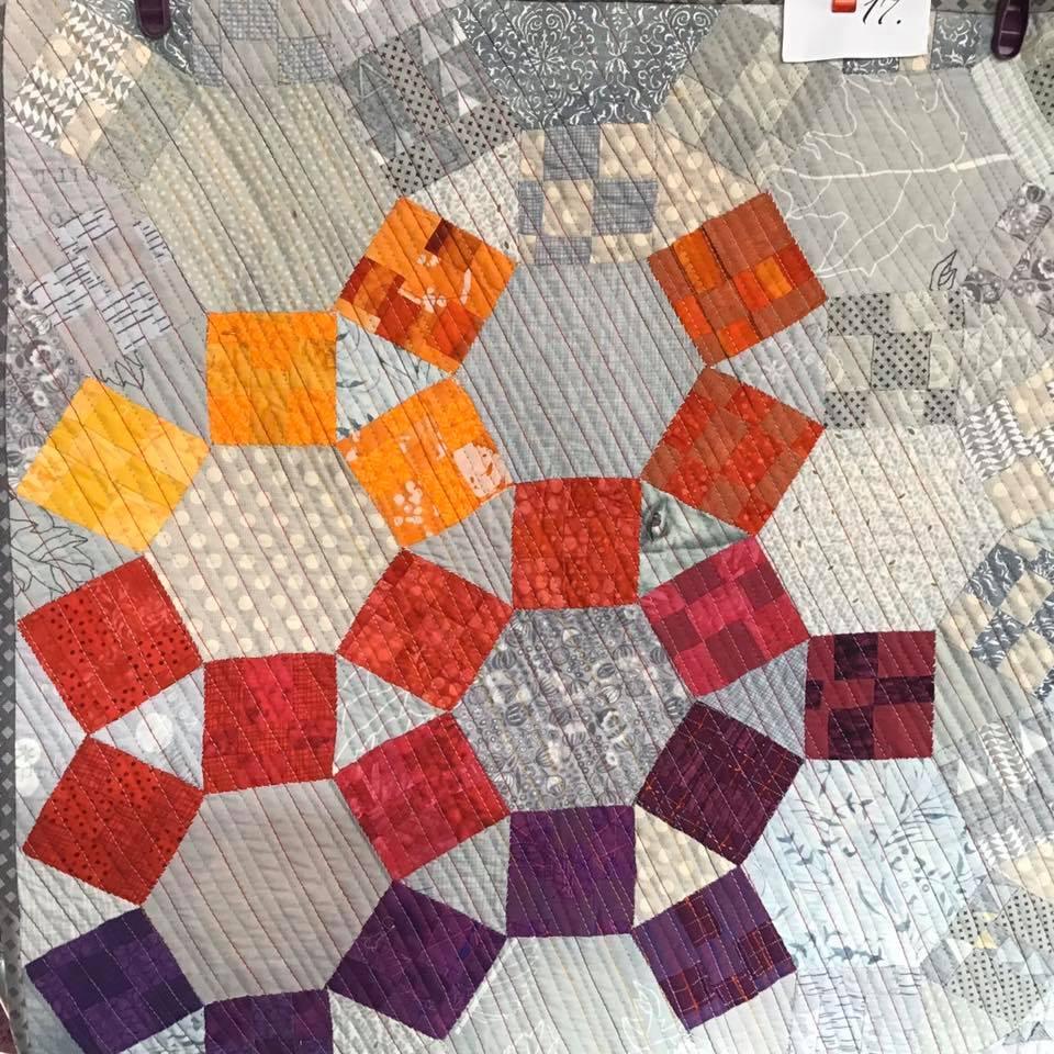 A small, square quilt with a grey background. The quilt features a hexagonal pattern of orange, red, and purple squares. The background is quilted with a diagonal line pattern.