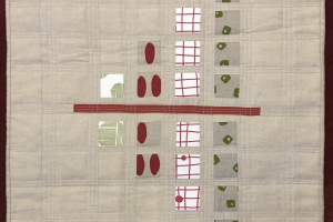 A quilt with a beige background. The quilt features a symmetrical design with red squares, green shapes, and white squares arranged in a grid-like pattern. A horizontal red line divides the quilt in half. The background is quilted with a subtle grid patte