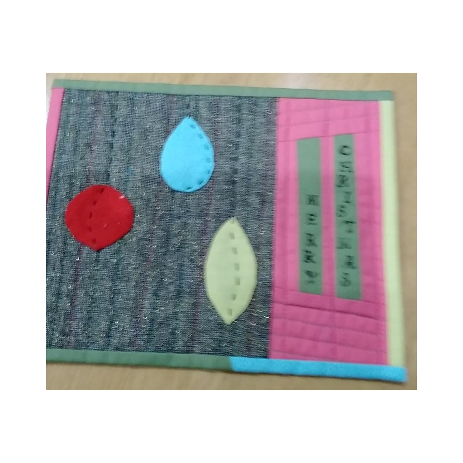 A rectangular quilt with a gray background. The quilt features three colored shapes: a red circle, a blue teardrop, and a yellow leaf. The shapes are sewn onto the gray background. There are also strips of pink, yellow, and blue fabric on the right side of the quilt.