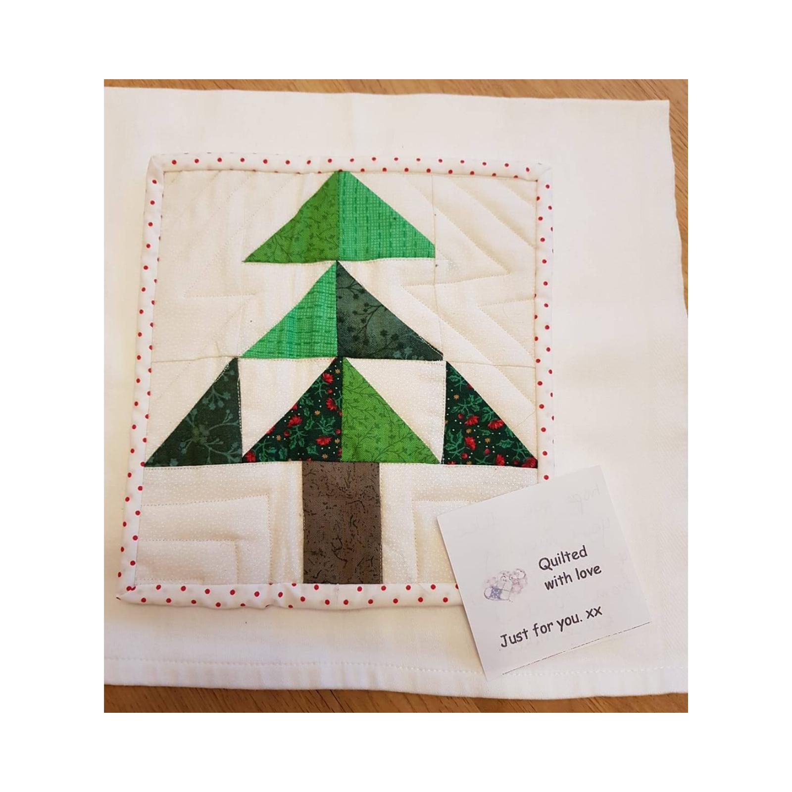 A square quilt with a white background and a red polka dot border. The quilt features a green Christmas tree made up of smaller triangles, with a brown trunk. A small tag attached to the quilt reads "Quilted with love. Just for you. xx"