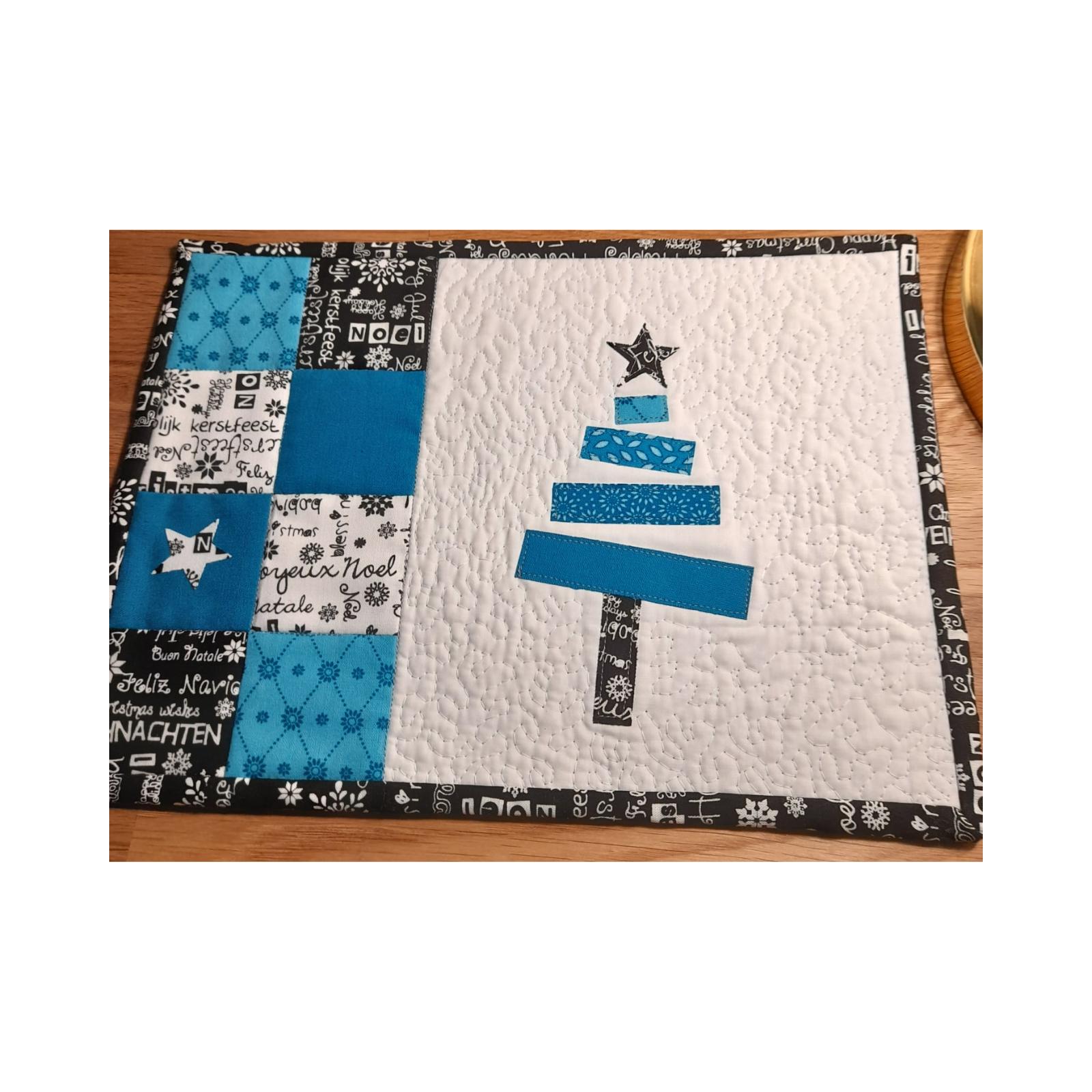 A rectangular quilt with a white background and a black border. The quilt features a blue Christmas tree made up of smaller blue squares. The background is filled with various Christmas greetings in different languages, including "Joyeux No&euml;l," "Frohe Weihnachten," and "Feliz Navidad."