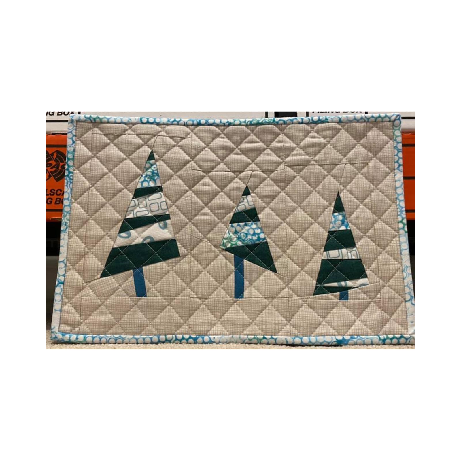 A rectangular quilt with a blue border. The quilt features three Christmas trees made of green and blue fabric with a pattern of horizontal lines. The trees are on a beige background, which is quilted with a diamond pattern.