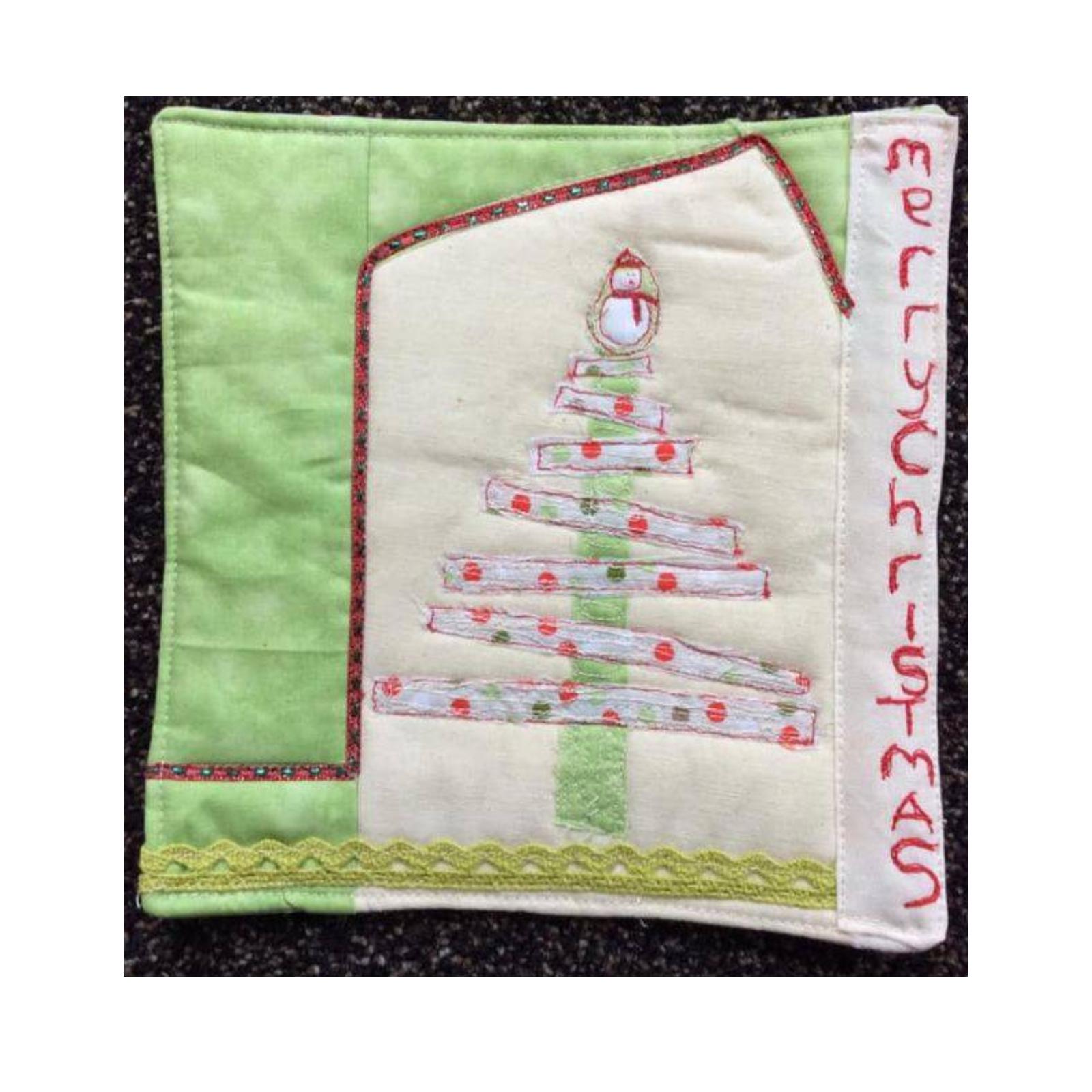 A square quilt with a green background and a white center section. The center section features a Christmas tree made of fabric strips with a snowman on top. The words "Merry Christmas" are embroidered on the right side of the quilt.