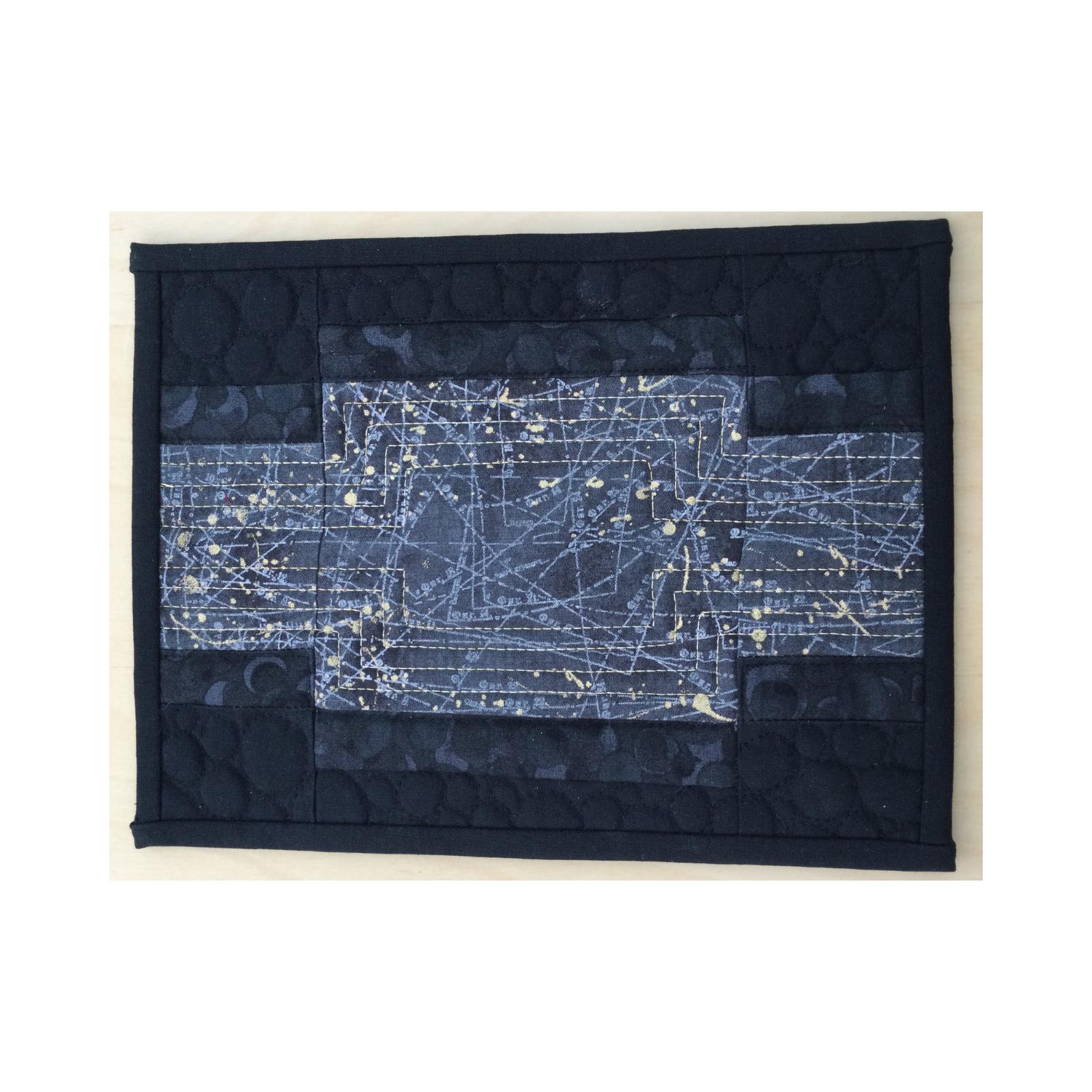 A rectangular quilt with a black background. The quilt features a central panel with a map of constellations, surrounded by black fabric with a pattern of small circles. The background is quilted with a subtle, linear pattern.