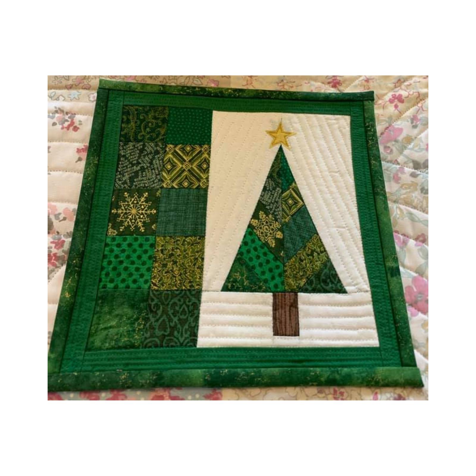 A square quilt with a green border. The quilt features a Christmas tree made up of green fabric triangles, with a brown trunk and a gold star on top. To the left of the tree is a patchwork of green fabric squares with various patterns. The background is a white fabric with a subtle, linear quilting pattern.