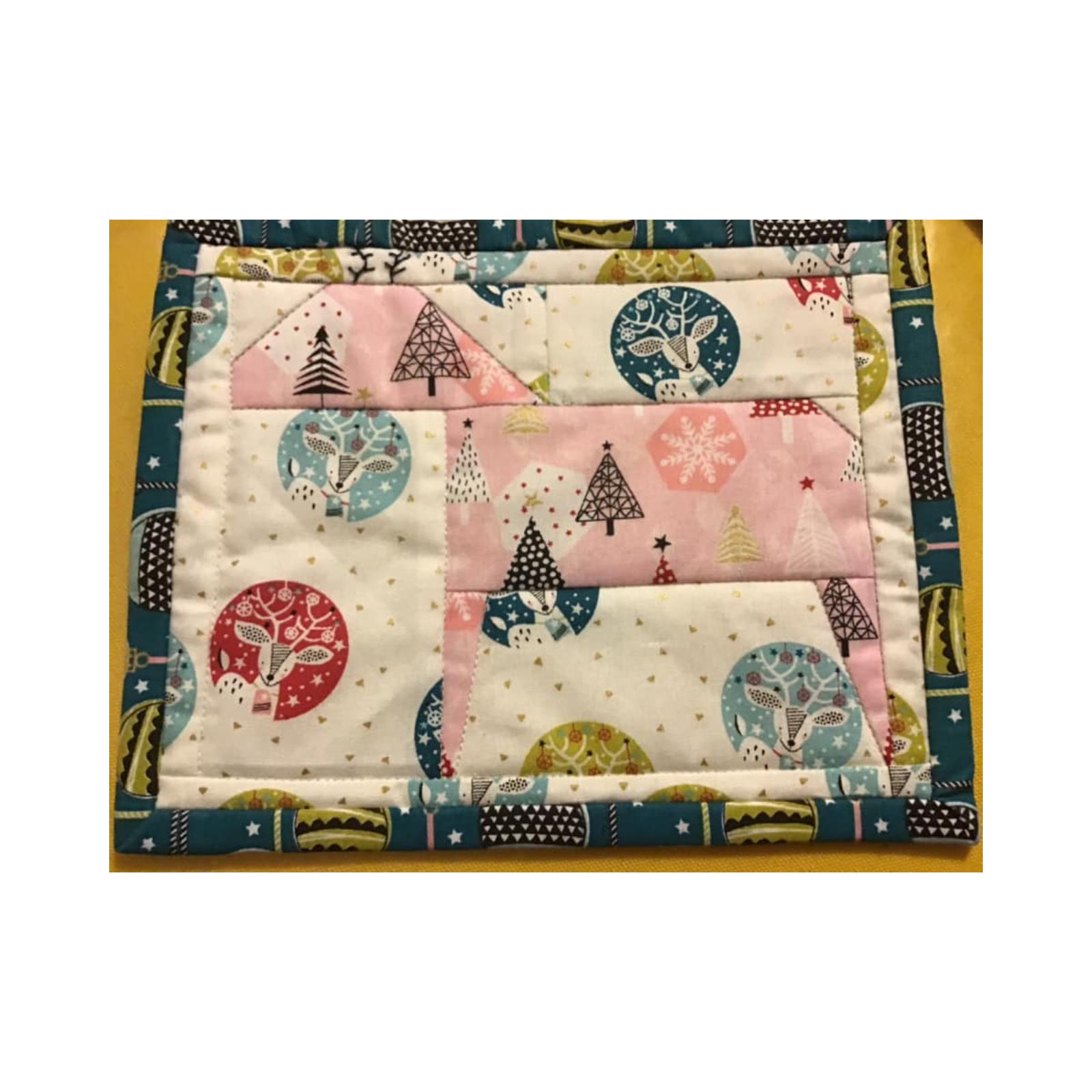 A rectangular quilt with a teal border. The quilt features a patchwork design with different fabrics, including pink, green, and gold. The fabrics have patterns of deer, trees, and snowflakes.