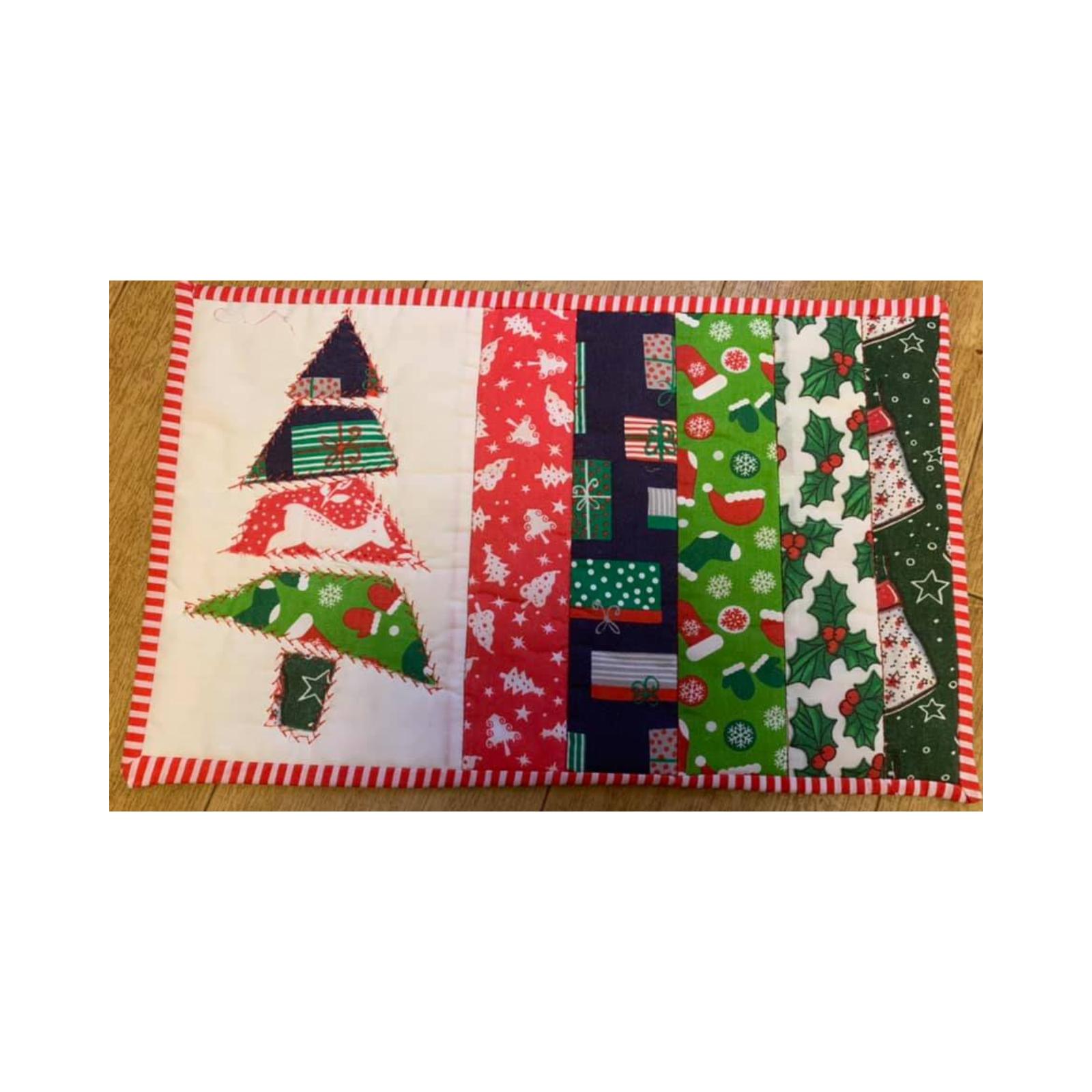 A rectangular quilt with a red border. The quilt features a green Christmas tree made up of patchwork squares on the left side. On the right side, there are vertical strips of fabric with Christmas-themed patterns, including presents, holly leaves, and snowflakes.
