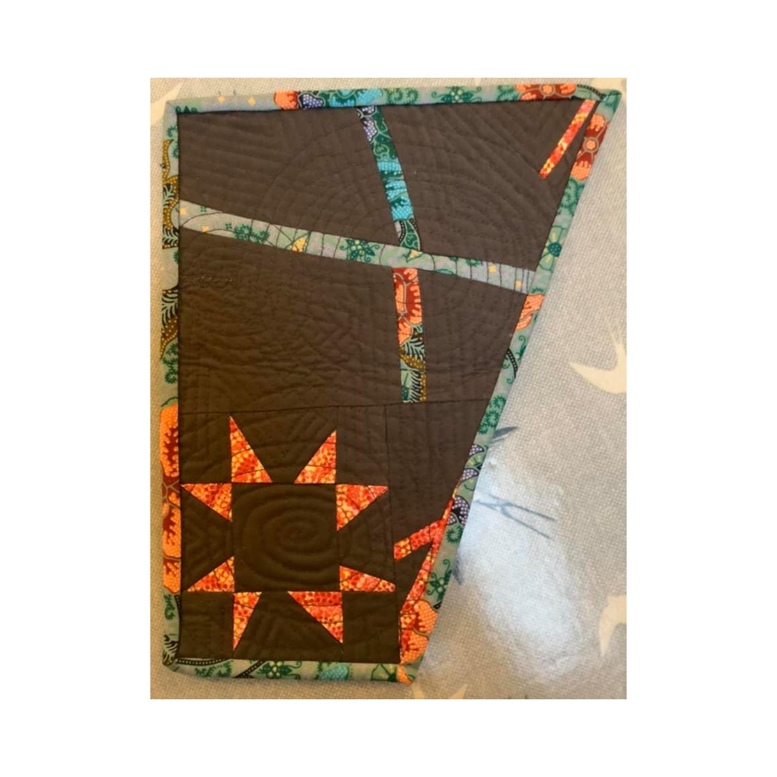 An irregular rectangle quilt with a brown background. The quilt features a patchwork design with orange and pink triangles in the bottom left corner. The rest of the quilt is a mix of brown fabric with lines and shapes cut out. The quilt is surrounded by a blue and green border.