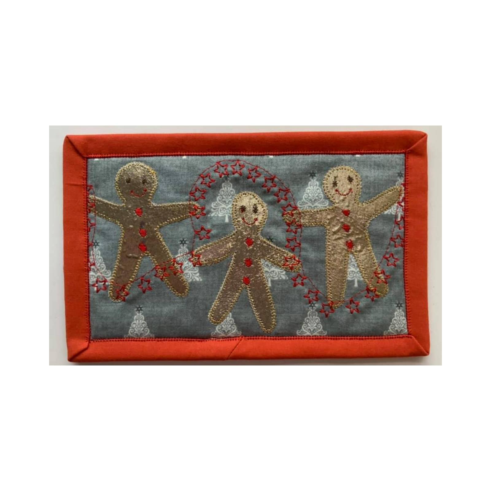 A rectangular quilt with a red border. The quilt features three gingerbread men holding hands and surrounded by a string of red stars. The background is a light grey fabric with a pattern of small Christmas trees.