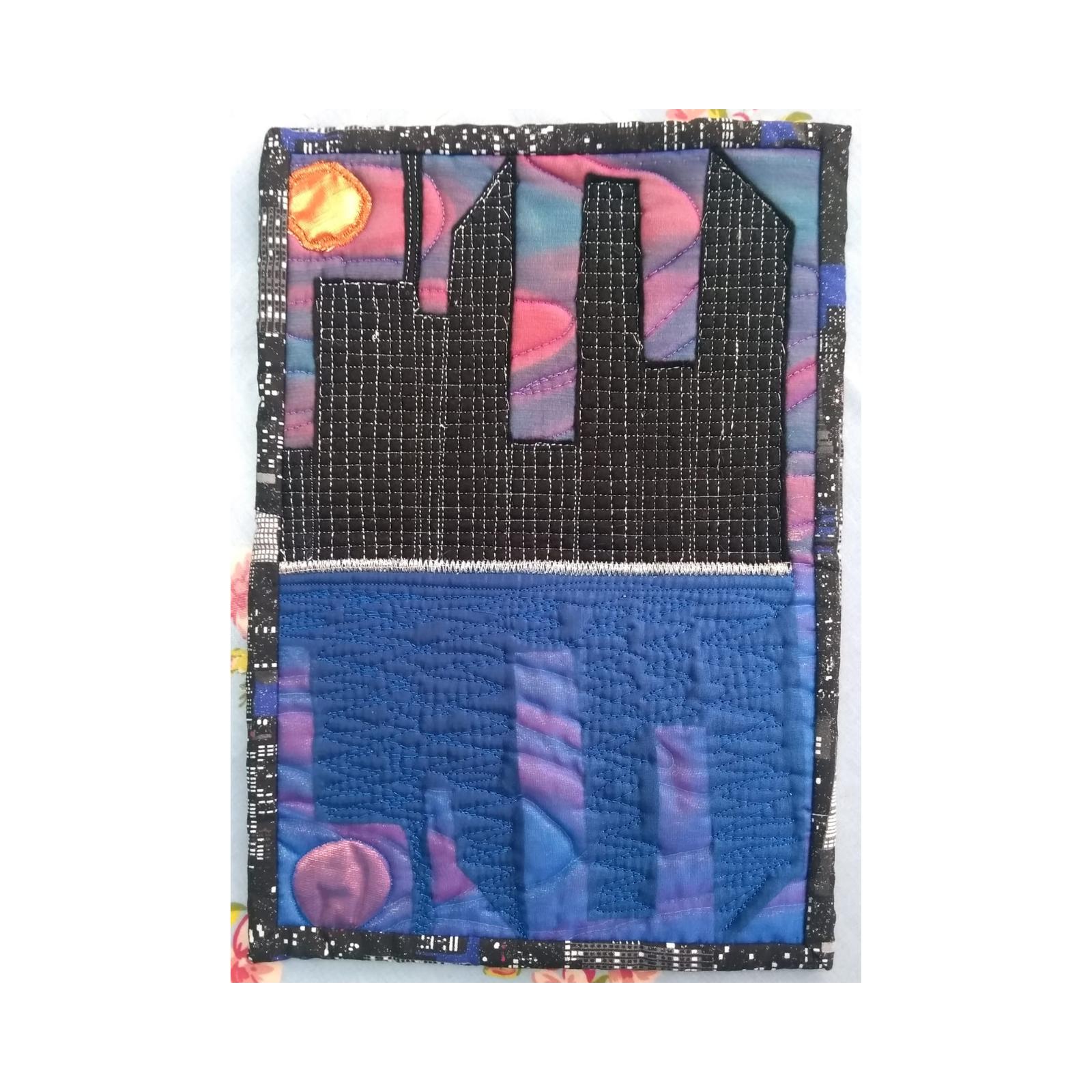 A square quilt with a black border. The quilt is divided into two sections. The top section shows a black silhouette of a city skyline against a yellow and purple sky. The bottom section is a dark blue background with a textured pattern representing a city at night.