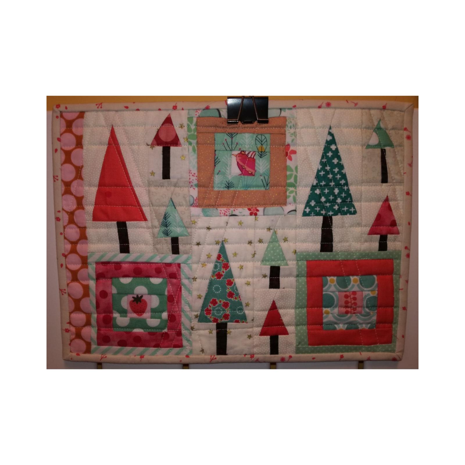 A rectangular quilt with a patchwork design. The quilt features multiple Christmas trees of various sizes and colors, including red, green, and pink. The trees are surrounded by squares of fabric with different patterns, including polka dots and stripes. The quilt is hanging from a clip.