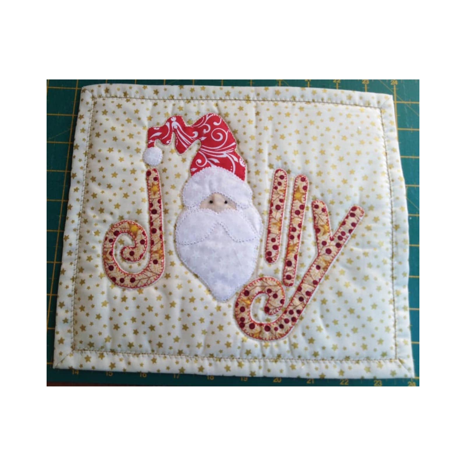 A rectangular quilt with a gold background and a green border. The word "JOLLY" is spelled out in colorful letters, with a small Santa Claus face in the centre of the "O." The background is filled with small gold stars.