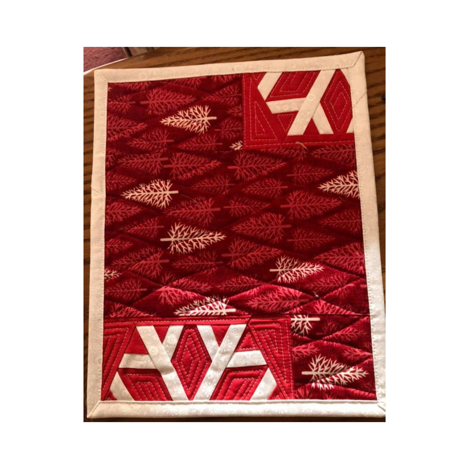 A rectangular quilt with a red background and a white border. The quilt features a white snowflake design in the centre, surrounded by a pattern of white trees on a red background. The background is quilted with a wavy pattern.