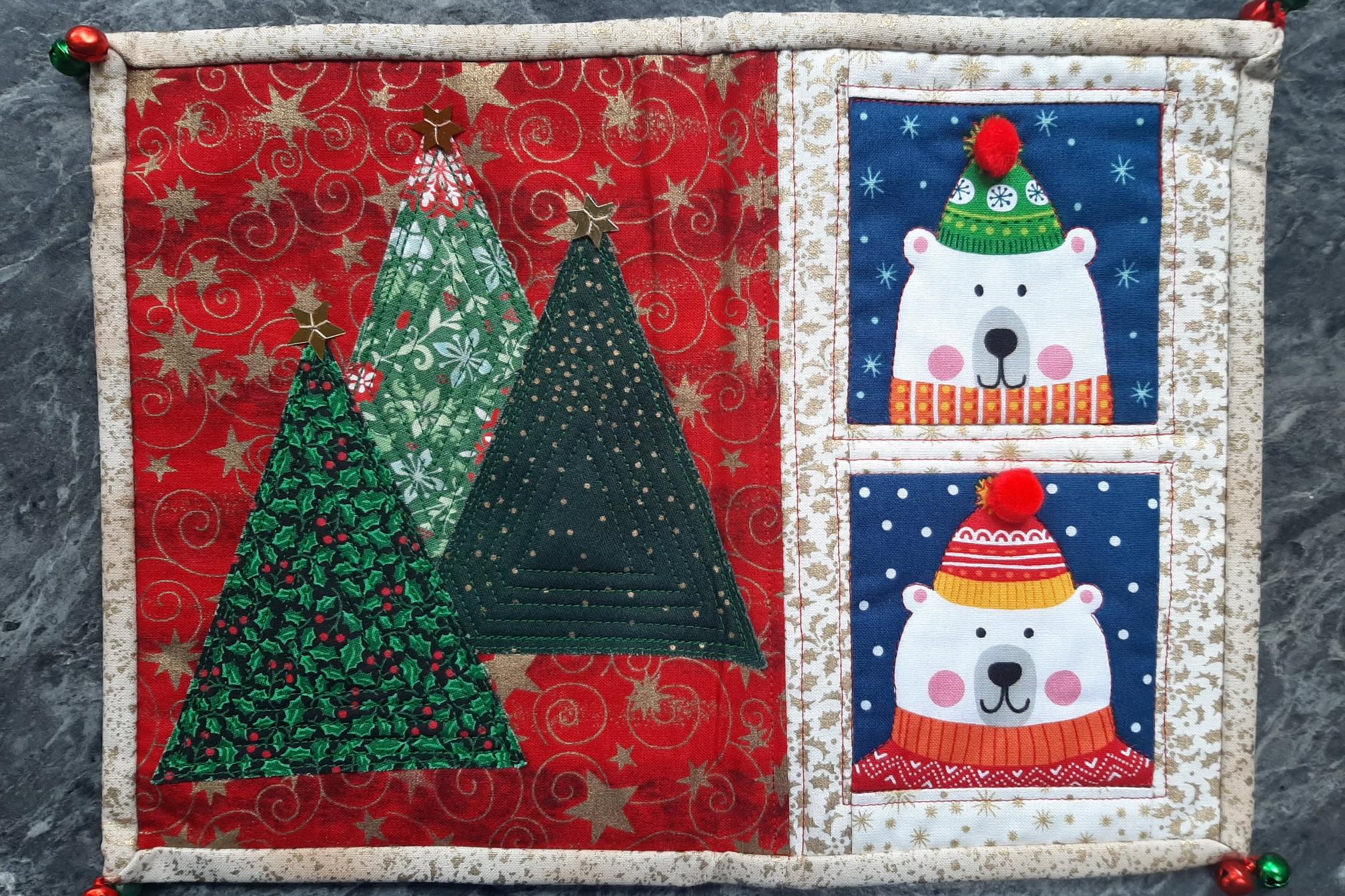 A rectangular quilt with a red background and a white border. The quilt is divided into two sections. The left section features two Christmas trees with ornaments and stars, while the right section shows two polar bears wearing hats and scarves, peeking o