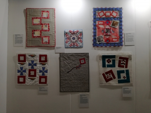 6 square modern quilts displayed on an exhibition wall. 