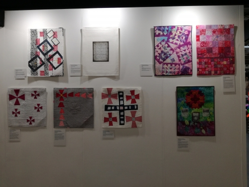 8 square modern quilts displayed on an exhibition wall. 