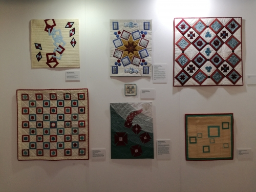 6 square modern quilts displayed on an exhibition wall. 