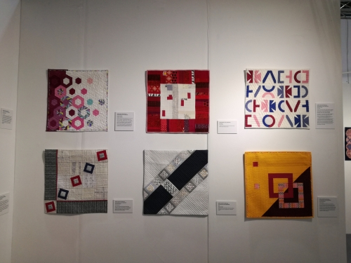 6 square modern quilts displayed on an exhibition wall. 