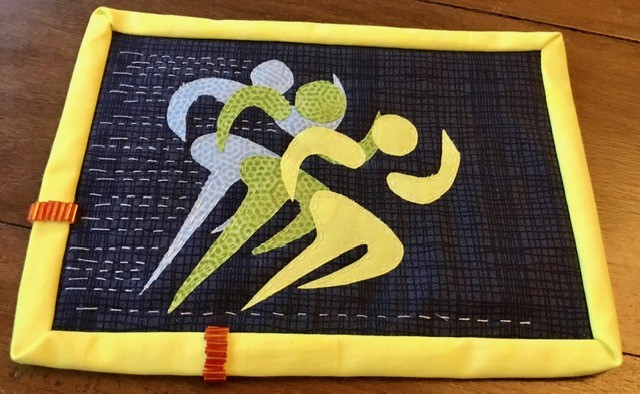 A postcard quilt with a yellow border. The quilt features three stylised figures running, with one in the foreground, one in the middle, and one in the background. The figures are in shades of yellow, green, and light blue, and the background is a hatched dark blue fabric. The quilt is quilted with a linear pattern. behind the runners to portray movement.