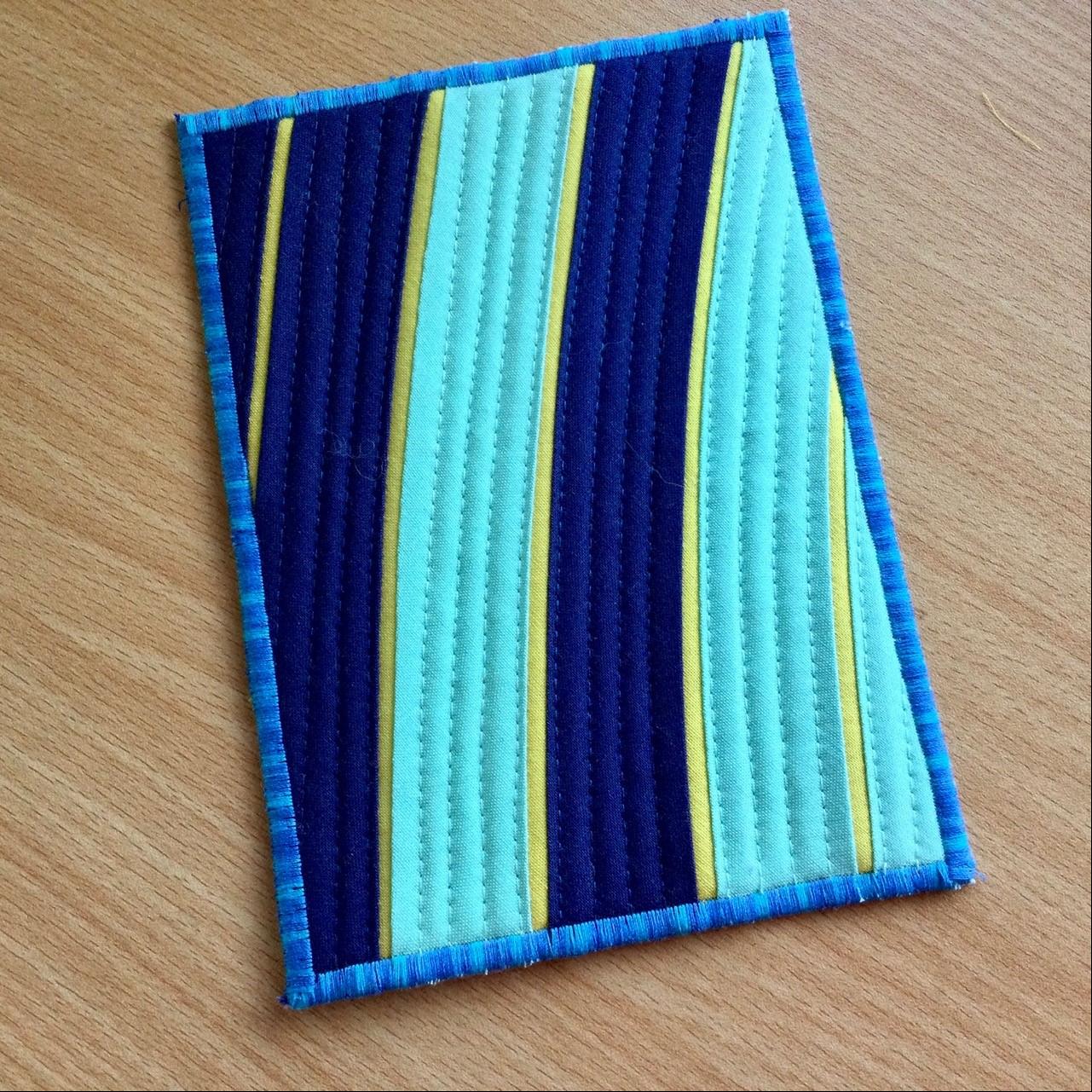A postcard quilt with a blue background and a blue border. The quilt features a pattern of vertical curving stripes in shades of blue and yellow. The stripes quilted following the curve of the stripe..