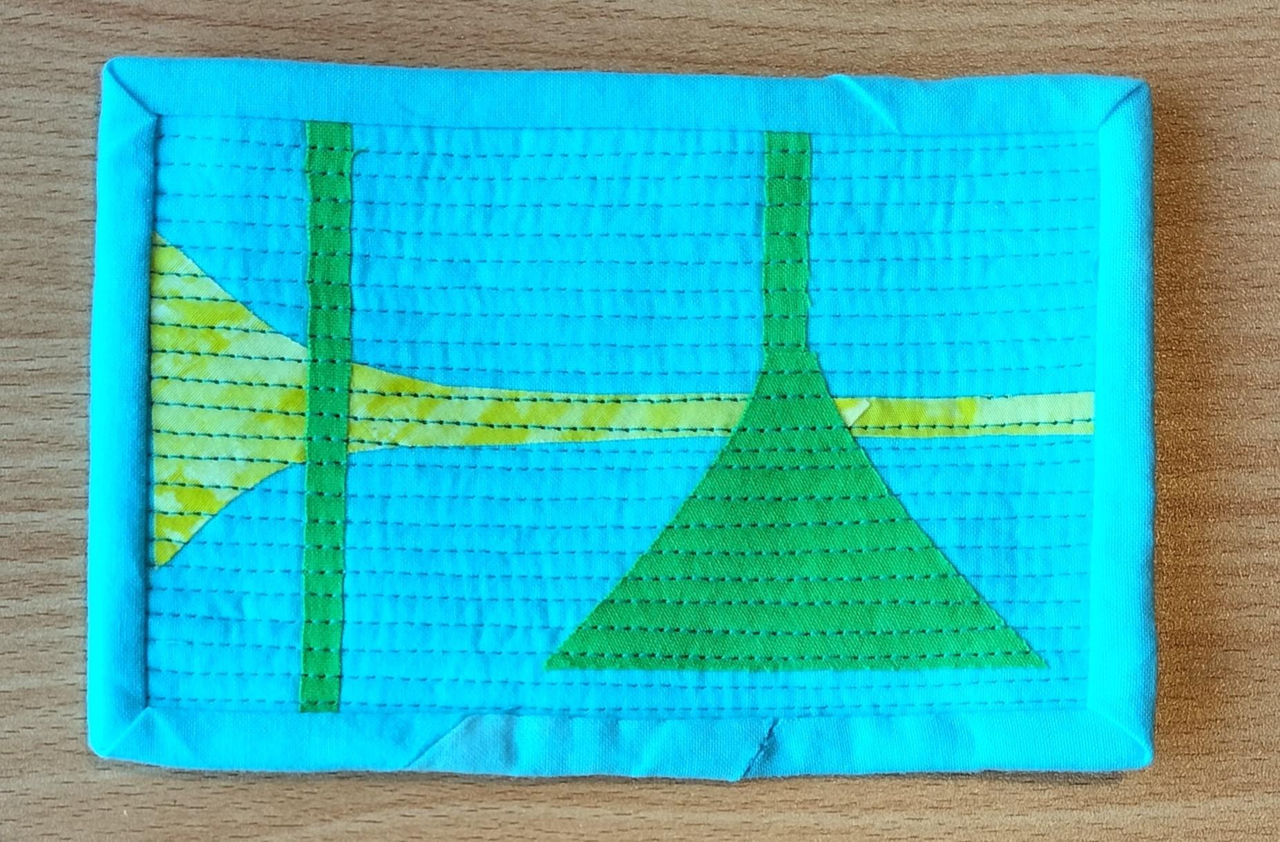 A postcard quilt with a blue background and a blue border. The quilt features a simple, abstract design with a yellow triangle, two green triangles, and two vertical green stripes. The background is quilted with horizontal lines.
