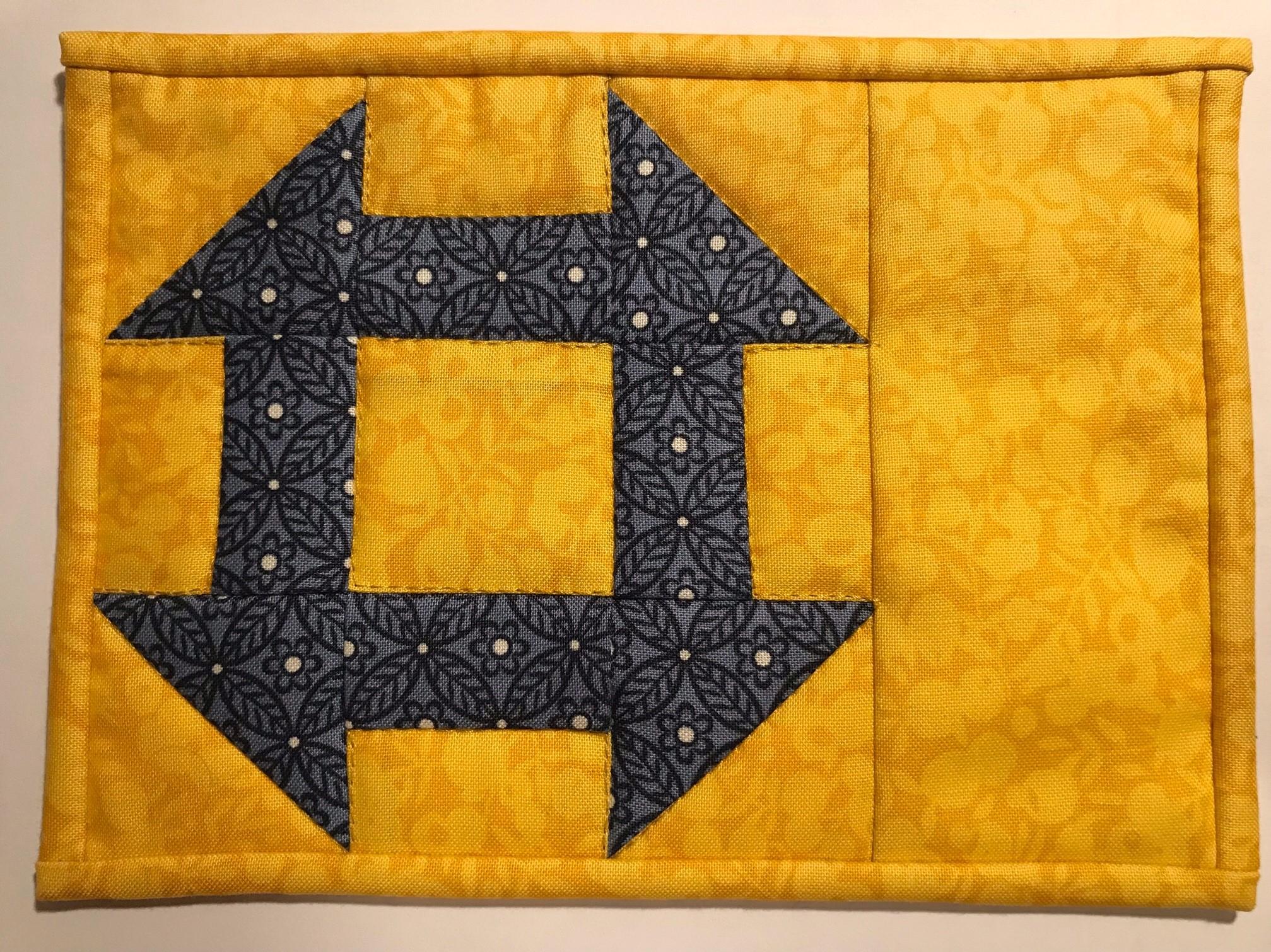 A postcard quilt with a yellow background. The quilt features churn dash design, made up of blue fabric with black leaves and flowers with yellow centres.