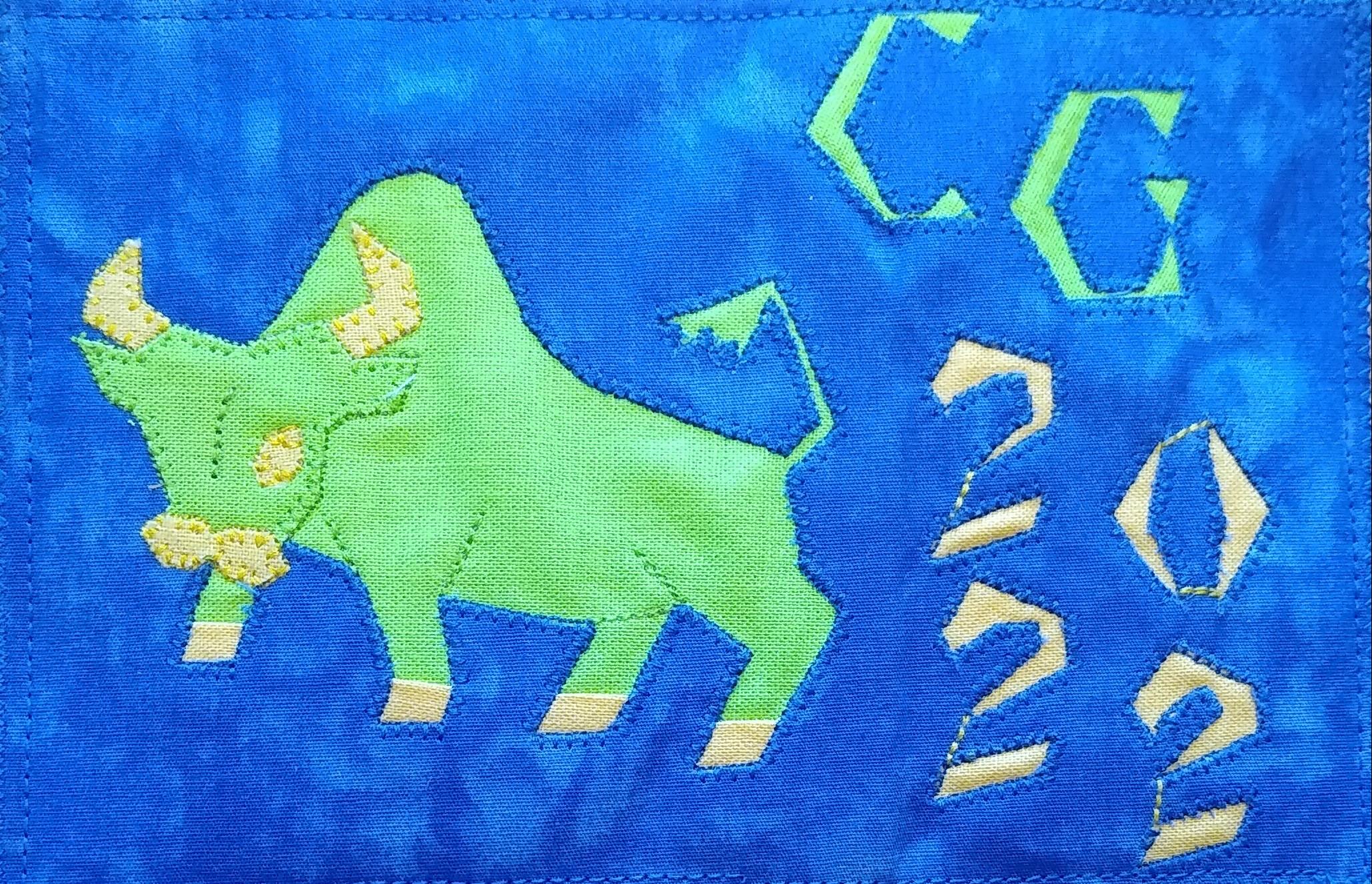 A postcard quilt with a blue background. The quilt features a green bull with yellow horns and hooves, standing on a blue field. The year "2022" is written in yellow letters in the lower right corner, and the letters "CG" are written in green letters in the top right corner.