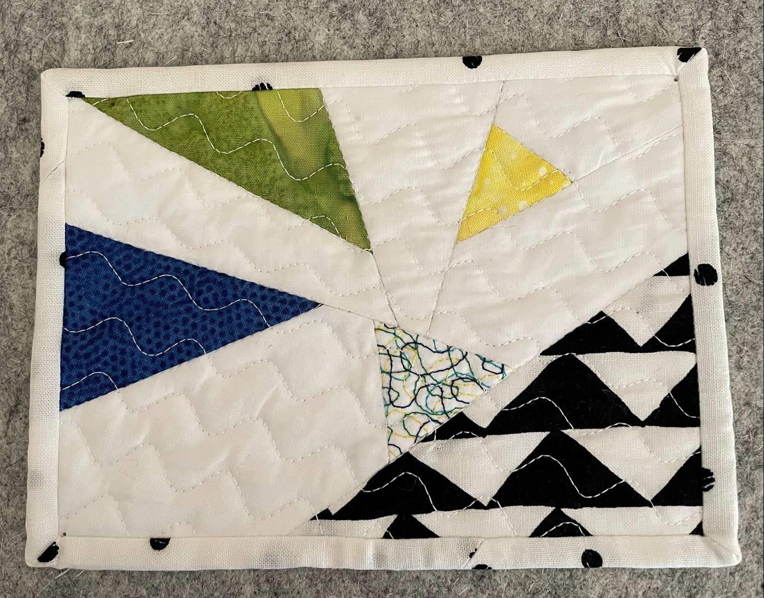 A postcard quilt with a white background and a black spotted border. The quilt features a colourful, abstract design with triangles in various colours and patterns, including green, yellow, blue, and black. The background is quilted with a wavy line pattern.