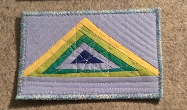 A postcard quilt with a grey background and a variegated stitched border. The quilt features a large triangle made up of smaller triangles in shades of blue, green, and yellow. The background is quilted with diagonal lines.