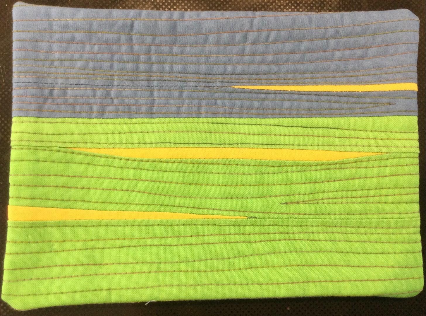 A postcard quilt with a green background and a blue-grey top section. The quilt features horizontal stripes of yellow fabric, creating a landscape-like scene with a sky and a field. The background is quilted with horizontal lines.