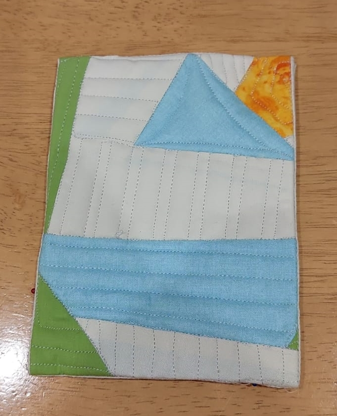 A postcard quilt with a white background. The quilt features a geometric design with triangles in shades of blue, green, and orange. The background is quilted with straight lines running horizontally and vertically..