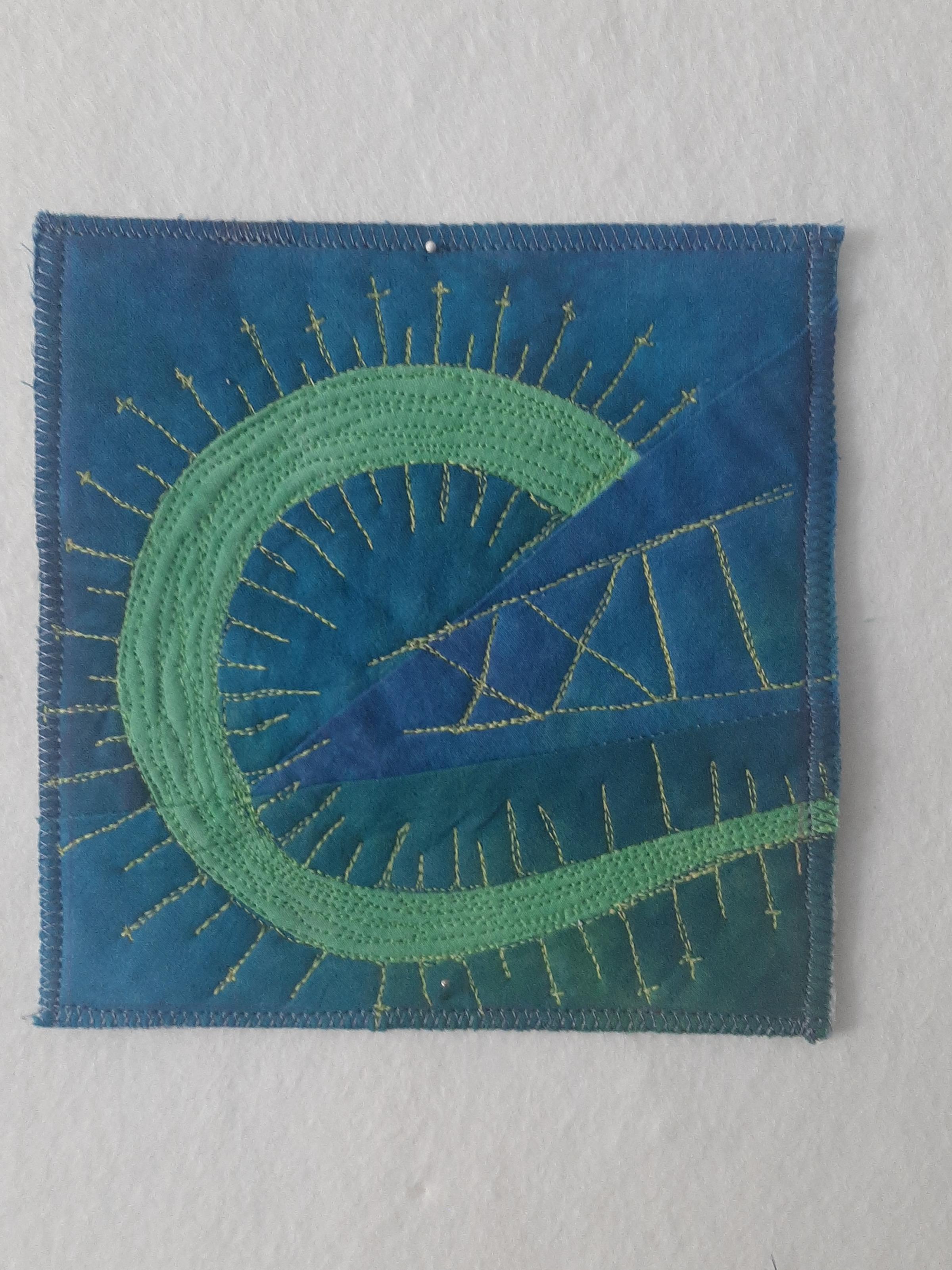 A postcard quilt with a pieced blue background. The quilt features a curved, green shape with a pointed end. The shape is surrounded by radiating lines of stitching. The roman numerals for the number 22 are stitched in the void of the C shape.