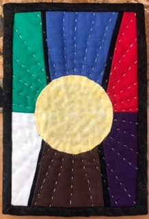 A postcard quilt with a black border. The quilt features a circular, yellow sun surrounded by colourful rays in red, blue, green, white, and brown. The background is quilted with a linear pattern radiating out from the central circle..