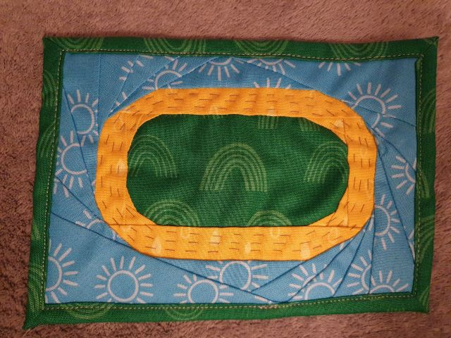 A rectangular postcard quilt with a blue background and a green border. The quilt features a circular, green patch surrounded by a yellow oval shape. The background is filled with a pattern of small, white suns