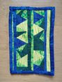 A postcard quilt with a blue background and a blue border. The quilt features a geometric design with triangles in shades of green and yellow. The triangles are arranged in a vertical pattern, creating a sense of movement. 