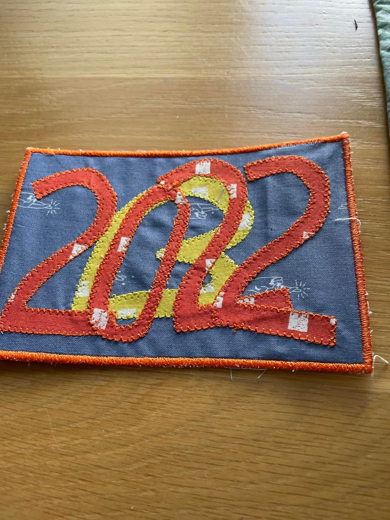 A rectangular postcard quilt with a blue background and an orange border. The quilt features the year "2022" written in orange and yellow fabric.