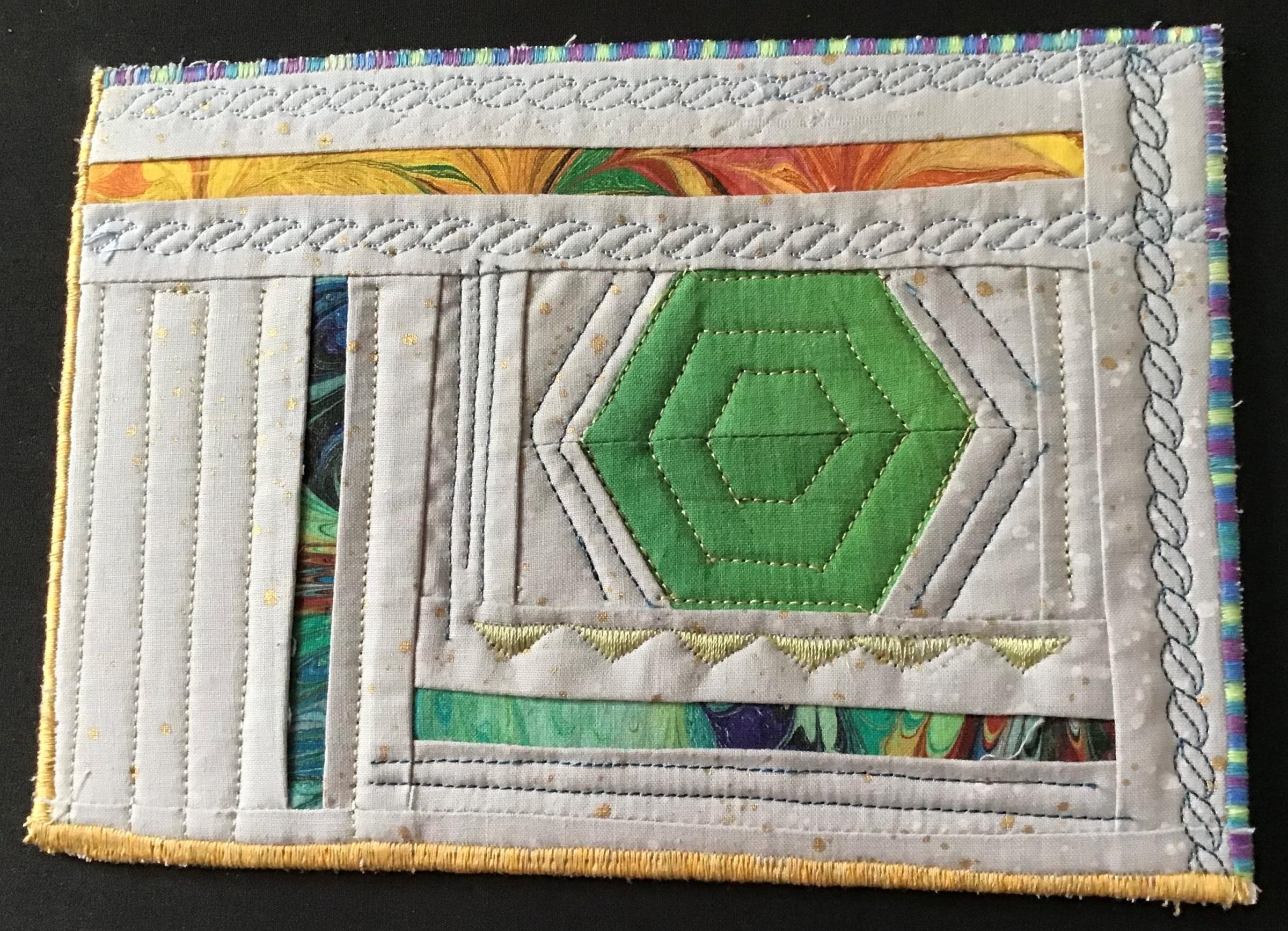 A postcard quilt with a light grey background and a colourful striped border. The quilt features a geometric design with a large, green hexagon in the centre, surrounded by various shapes and patterns in shades of blue, green, yellow, and orange. The background is quilted with different stitches and lines.