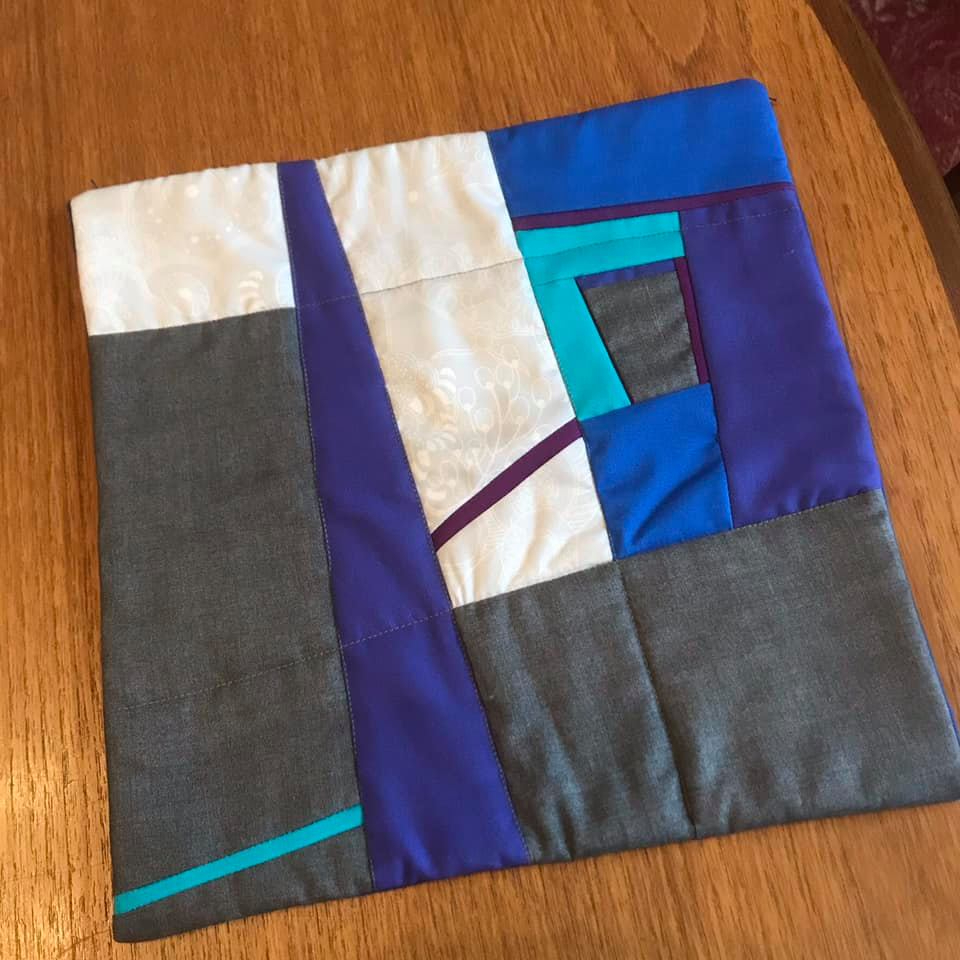 A small, square quilt with a patchwork design. It features various shades of blue, purple, gray, and white fabrics arranged in geometric shapes. There are also thin strips of teal fabric adding a pop of colour.