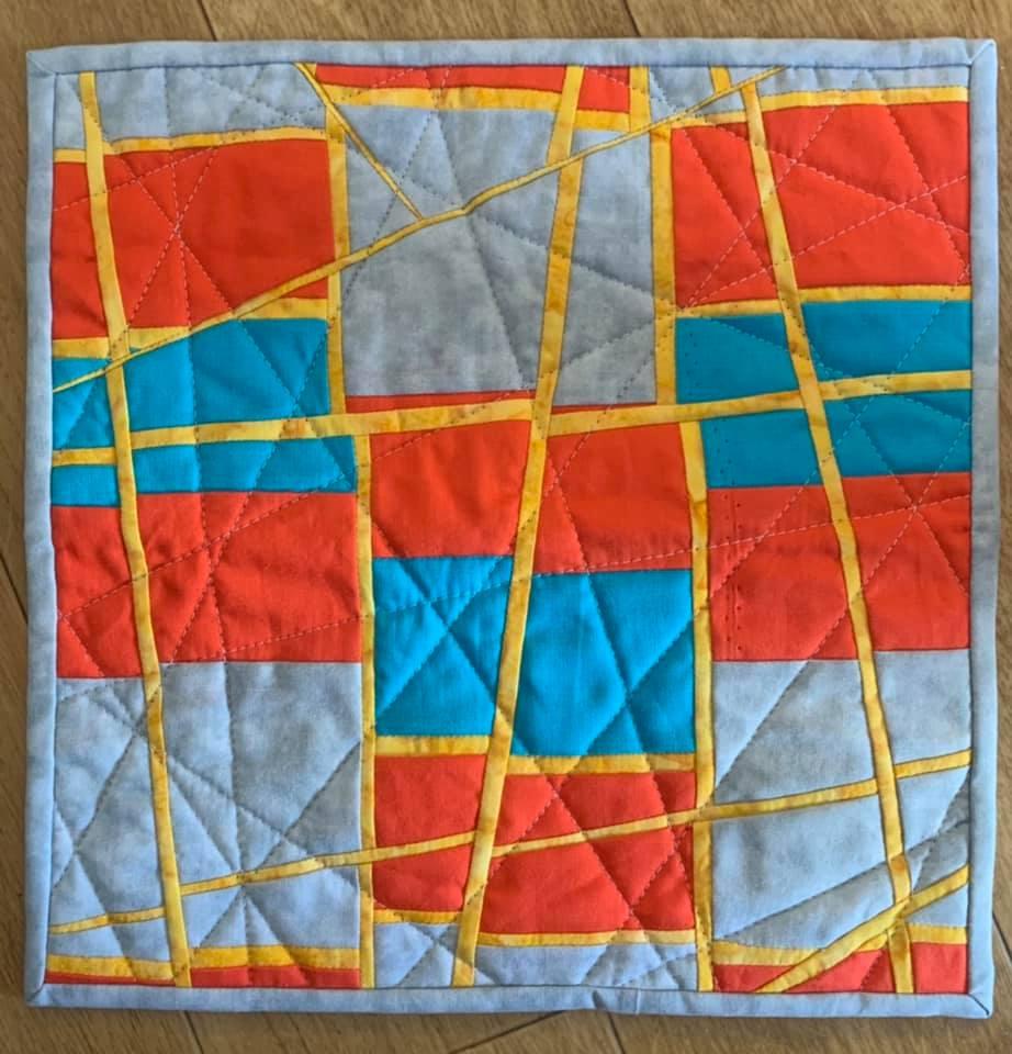 A small, square quilt with a patchwork design. It features a combination of red, blue, grey, and yellow fabrics arranged in geometric shapes and lines. The quilt has a textured, quilted pattern.