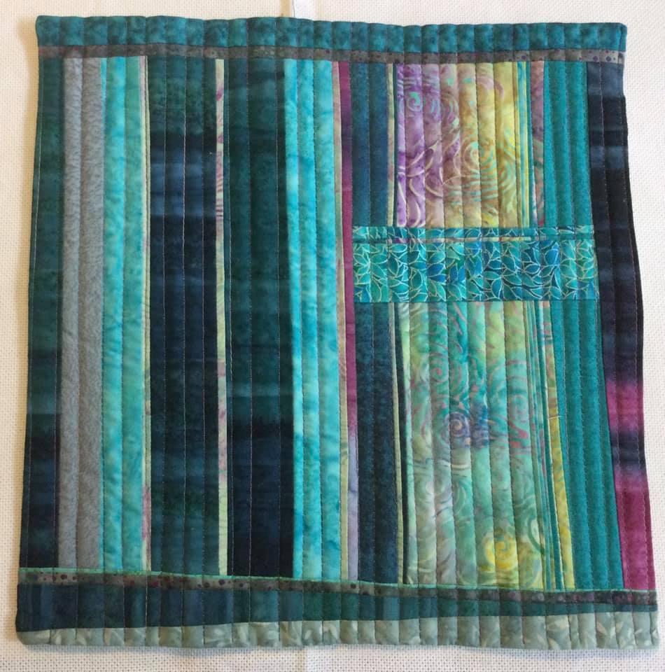 A small, rectangular quilt with a patchwork design. It features various shades of blue and green fabrics arranged in vertical strips. Some strips have a textured or patterned design.